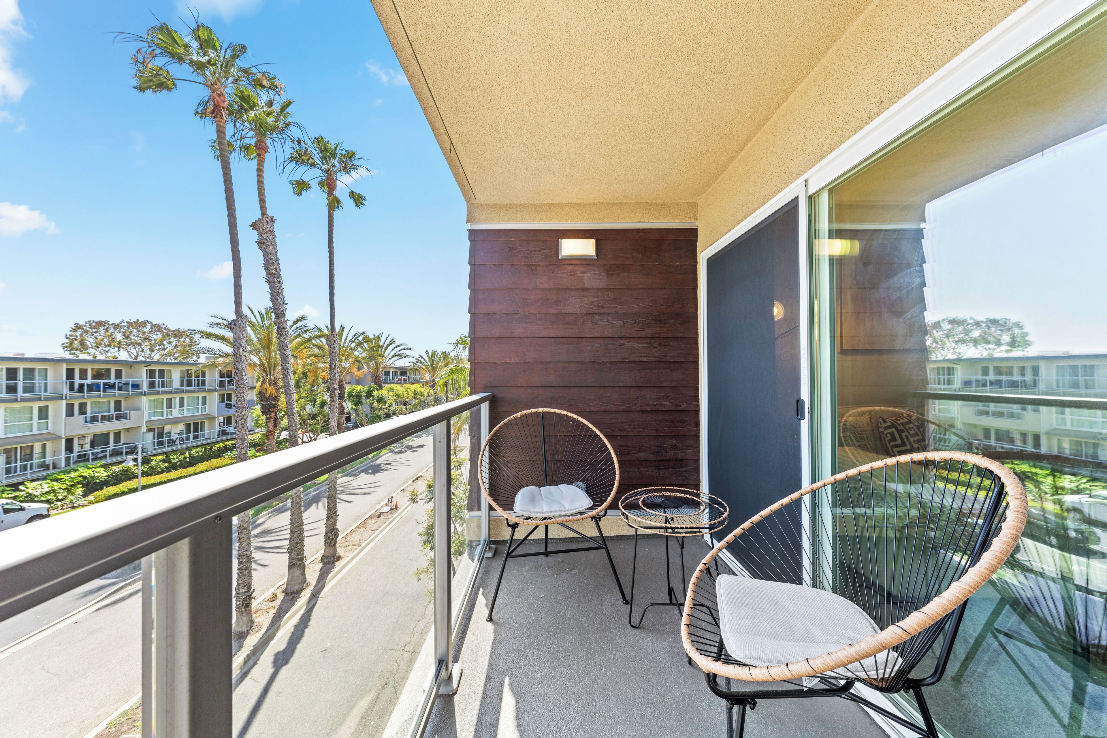A balcony with two wicker chairs, a small round table, and a view of palm trees and buildings.