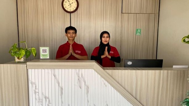 A reception desk with two individuals standing behind it, a clock above, and a plant to the left. - Bell Hotel Merauke (Merauke)
