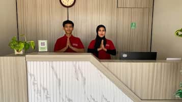 A reception desk with two individuals standing behind it, a clock above, and a plant to the left.