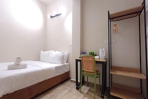 Una cama individual con ropa de cama blanca, un escritorio pequeño con una silla, un estante y una repisa en la pared.