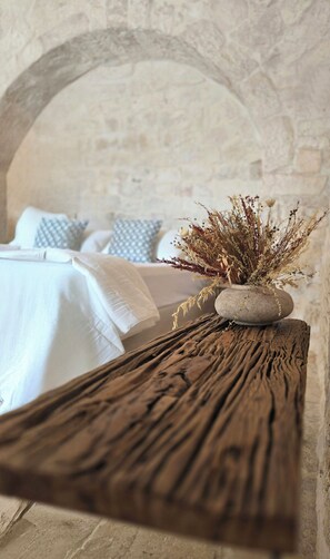 A rustic wooden table with a stone vase containing dried flowers, set against a stone wall with a bed and patterned pillows in the background. - Art-filled farmhouse in the Valle d'Itria trulli (Locorotondo)