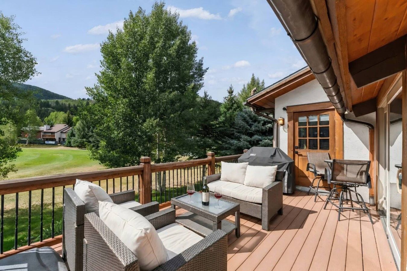 Terraza de madera con mobiliario de mimbre, una parrilla y vista a un campo de golf.