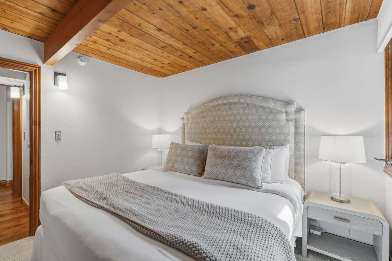Un dormitorio con techo de madera, una cama con almohadas estampadas, una lámpara de noche y una mesita de noche.