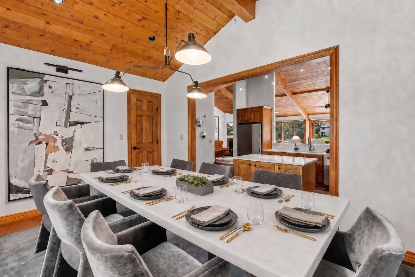 Comedor con una larga mesa dispuesta para seis comensales, vigas de madera en el techo y vista a la cocina.