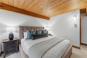 Un dormitorio con cabecera capitonada, cama con edredón blanco, manta gris y mesita de noche con lámpara.