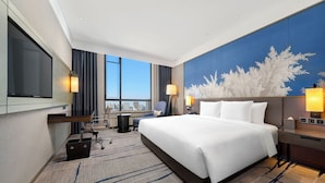 A hotel room with a large bed, a desk, a chair, a TV, and a view of the city. - Steigenberger Harbin Sunac Land (Harbin)