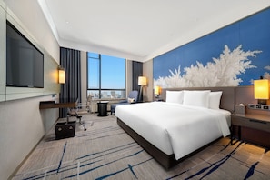 A hotel room with a large bed, a desk, a chair, a TV, and a view of the city. - Steigenberger Harbin Sunac Land (Harbin)