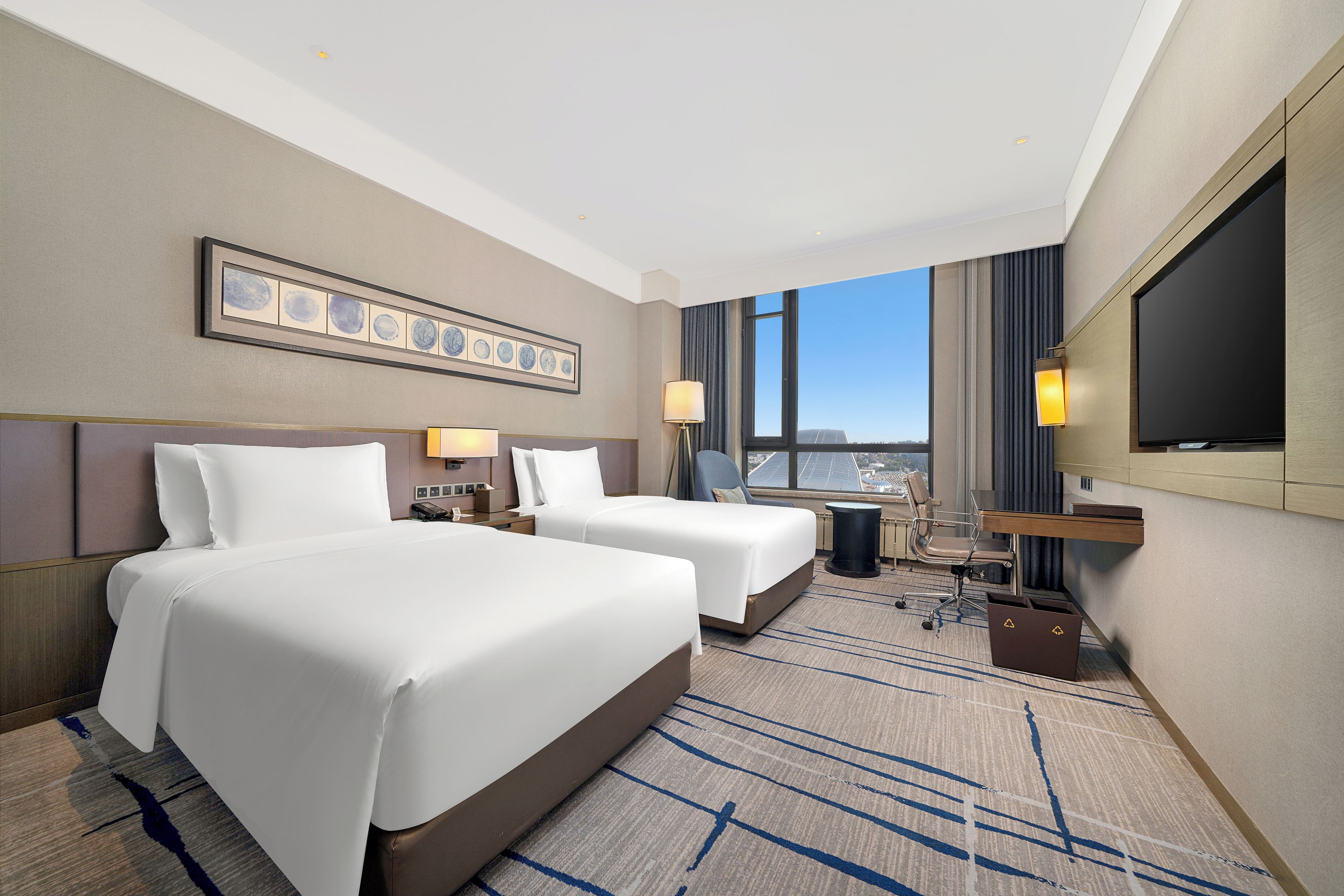 A hotel room with two beds, a desk, a chair, a TV, and a city view.