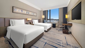 A hotel room with two beds, a desk, a chair, a TV, and a city view. - Steigenberger Harbin Sunac Land (Harbin)