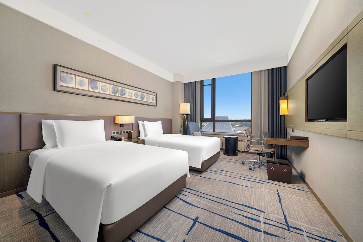 A hotel room with two beds, a desk, a chair, a TV, and a city view.