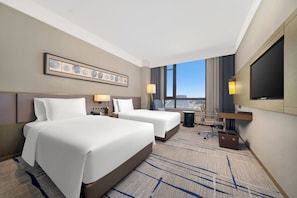 A hotel room with two beds, a desk, a chair, a TV, and a city view. - Steigenberger Harbin Sunac Land (Harbin)