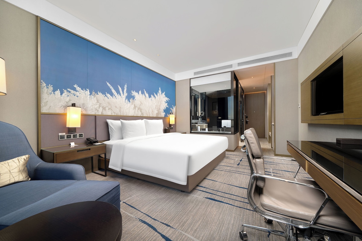 A hotel room with a large bed, a desk, a chair, a TV, and a bathroom visible through a glass door.
