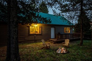 A cabin with a deck, two windows with lights, a door, and a fire pit with chairs. - Rustic Cabin with Firepit for Cozy Summer Night Escapes in Island Park, Idaho (Island Park)