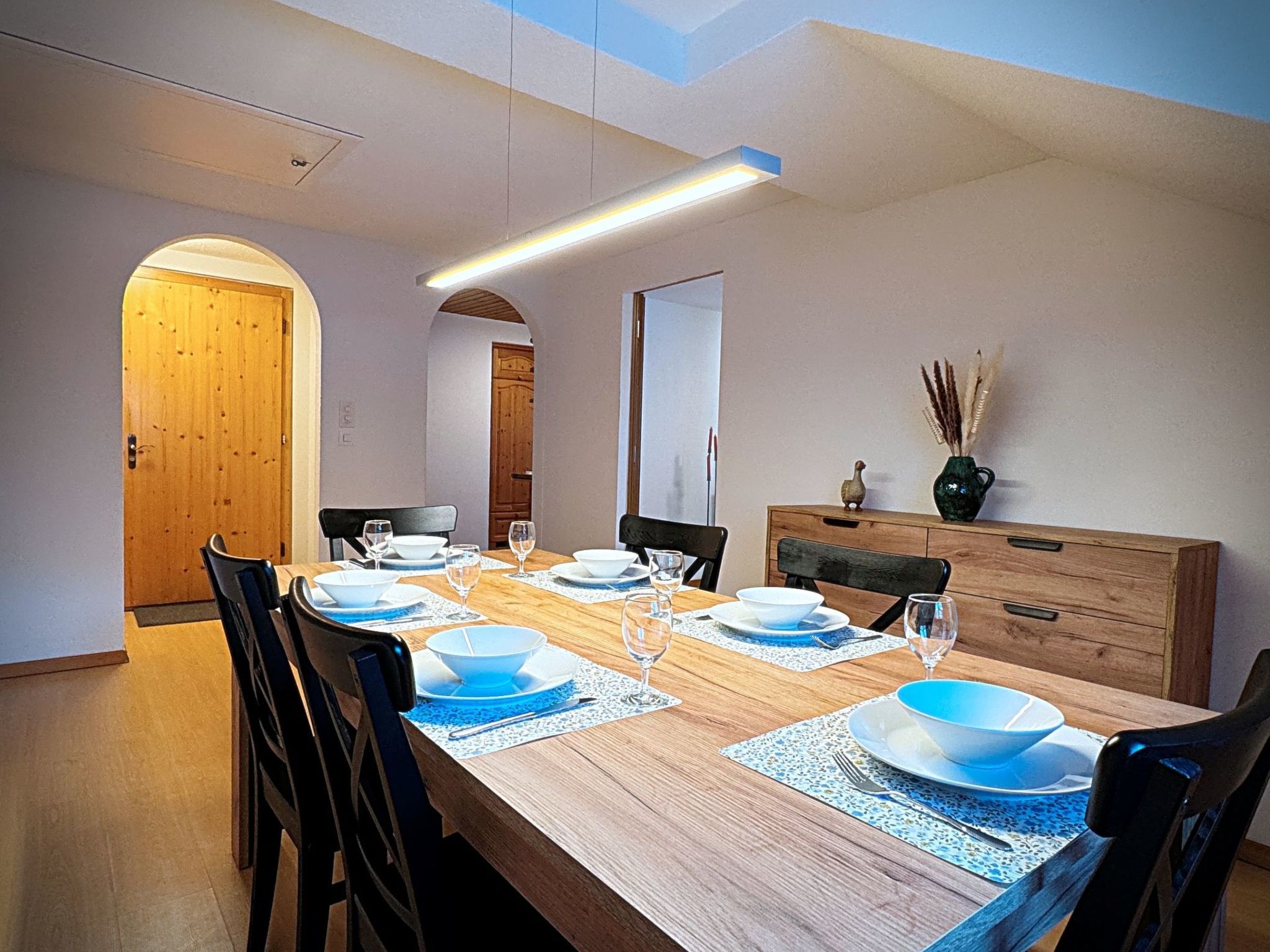 Une salle à manger avec une longue table en bois, dressée pour quatre personnes, des chaises et un buffet en bois.