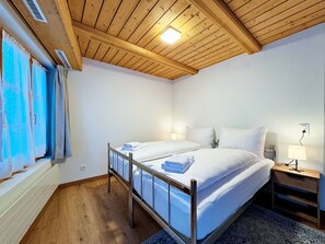 Une chambre avec un plafond en bois, un lit recouvert de draps blancs, une table de chevet avec une lampe et une fenêtre avec des rideaux.
