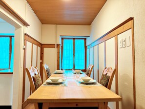 A dining room with a long wooden table set for four, flanked by chairs with patterned cushions, a window with curtains, and a wooden ceiling.