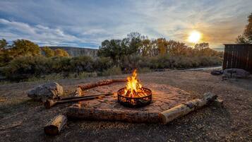 A campfire with logs and a metal fire pit in a natural setting with trees and a clear sky.