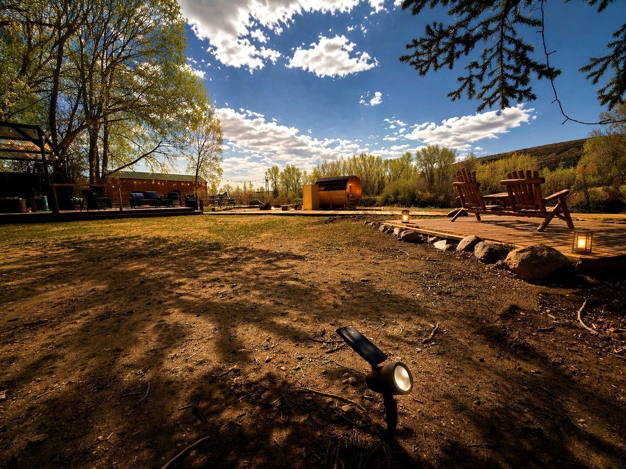 An outdoor area with a dirt ground, wooden chairs, a metal barrel, and a building in the background.