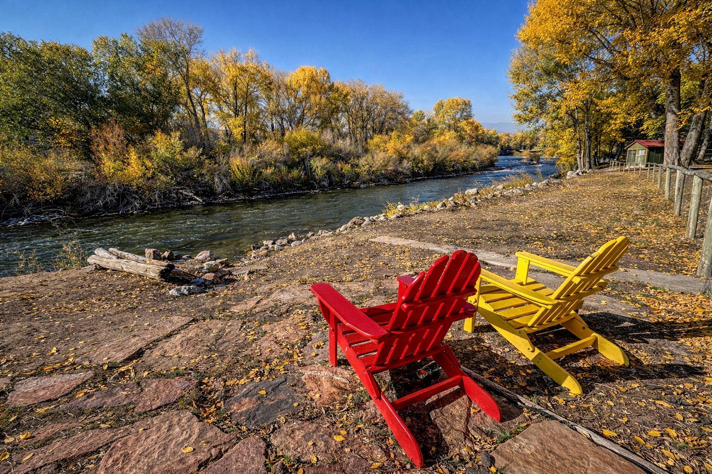 A riverside view with a wooden dock, a red and a yellow chair, and a small cabin in the background.