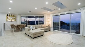 A modern living room with a sofa, chairs, a dining table, and a large sliding glass door leading to a balcony with a view. - Casa Rola Al Mar – Relax & Enjoy Stunning Ocean Views (Forster)