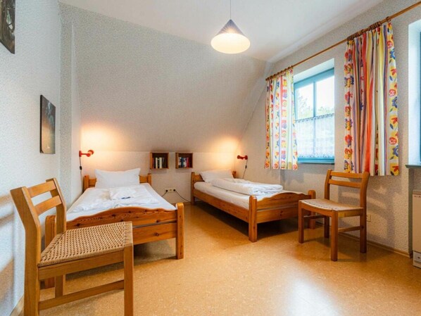 Ett rum med två sängar i trä, en stol i trä, ett litet bord och ett fönster med gardiner.
