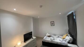 Un dormitorio con una cama, un televisor sobre un soporte, una mesita de noche e imagen colgada en la pared.