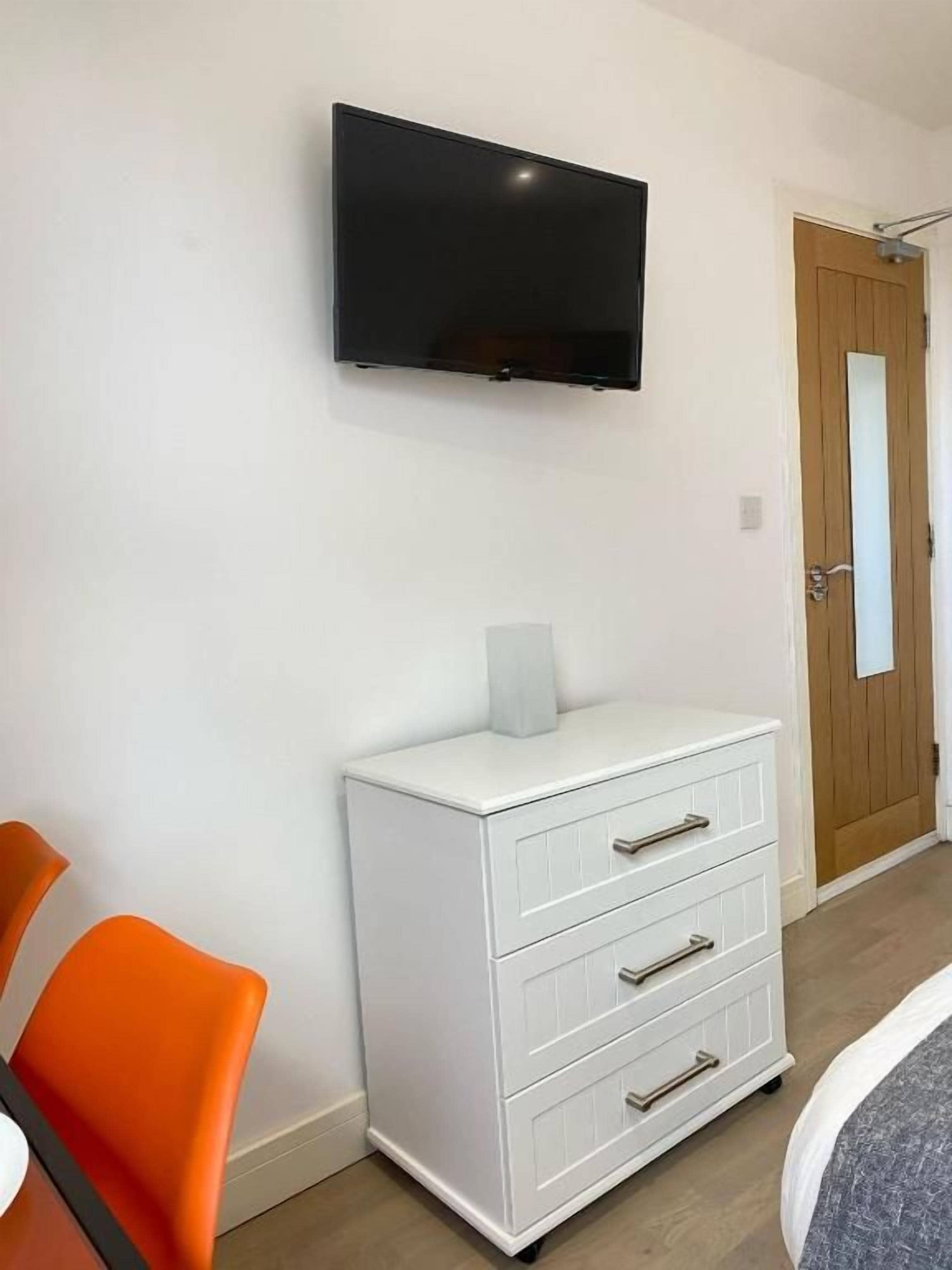 Une pièce dotée d’un téléviseur à écran plat fixé au mur, d’une commode blanche avec des tiroirs et d’une porte en bois.