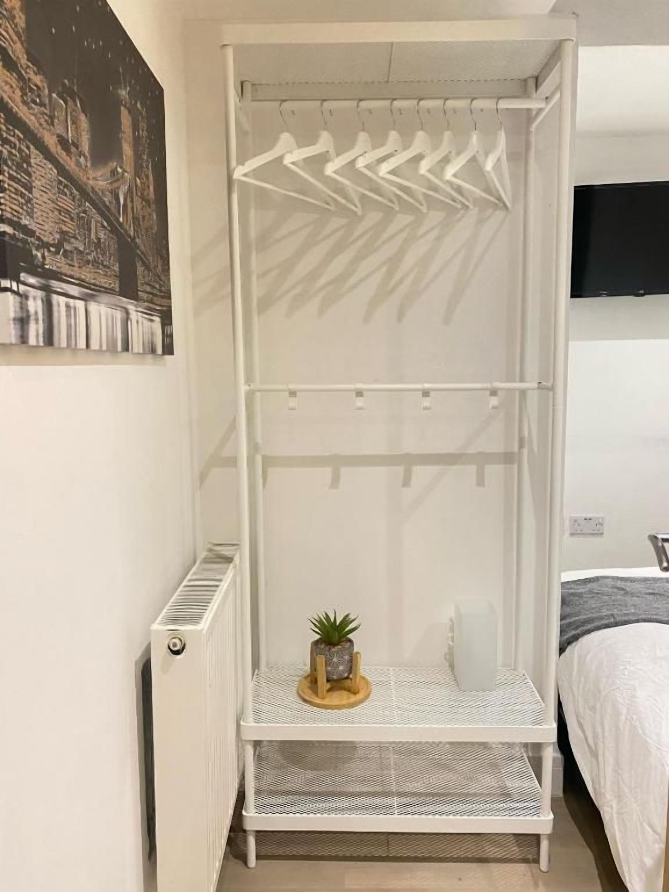 Un armario metálico blanco con barra para colgar y un estante, una planta pequeña sobre el estante, y un radiador en la pared.