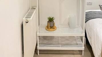 Un armario metálico blanco con barra para colgar y un estante, una planta pequeña sobre el estante, y un radiador en la pared.