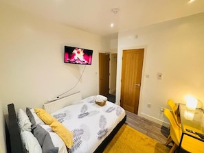 A bedroom with a bed, a television mounted on the wall, a wooden door, and a small table with a chair. - Stratford Green Nest (Greater London)
