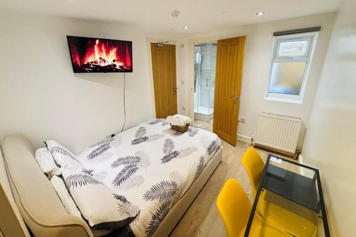 Un dormitorio con cama, un televisor que muestra una chimenea, una ventana y una puerta que da a otra habitación.