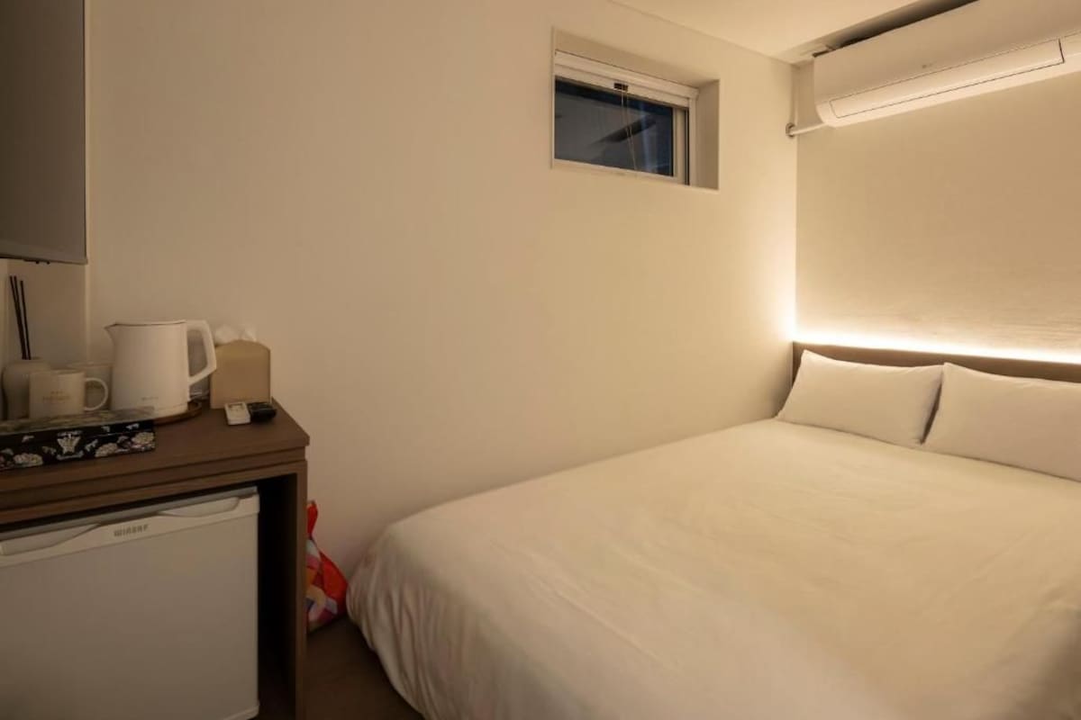 Quarto de hotel com cama, uma mesinha com chaleira, chávenas e uma caixa, uma janela e um ar condicionado fixado na parede.