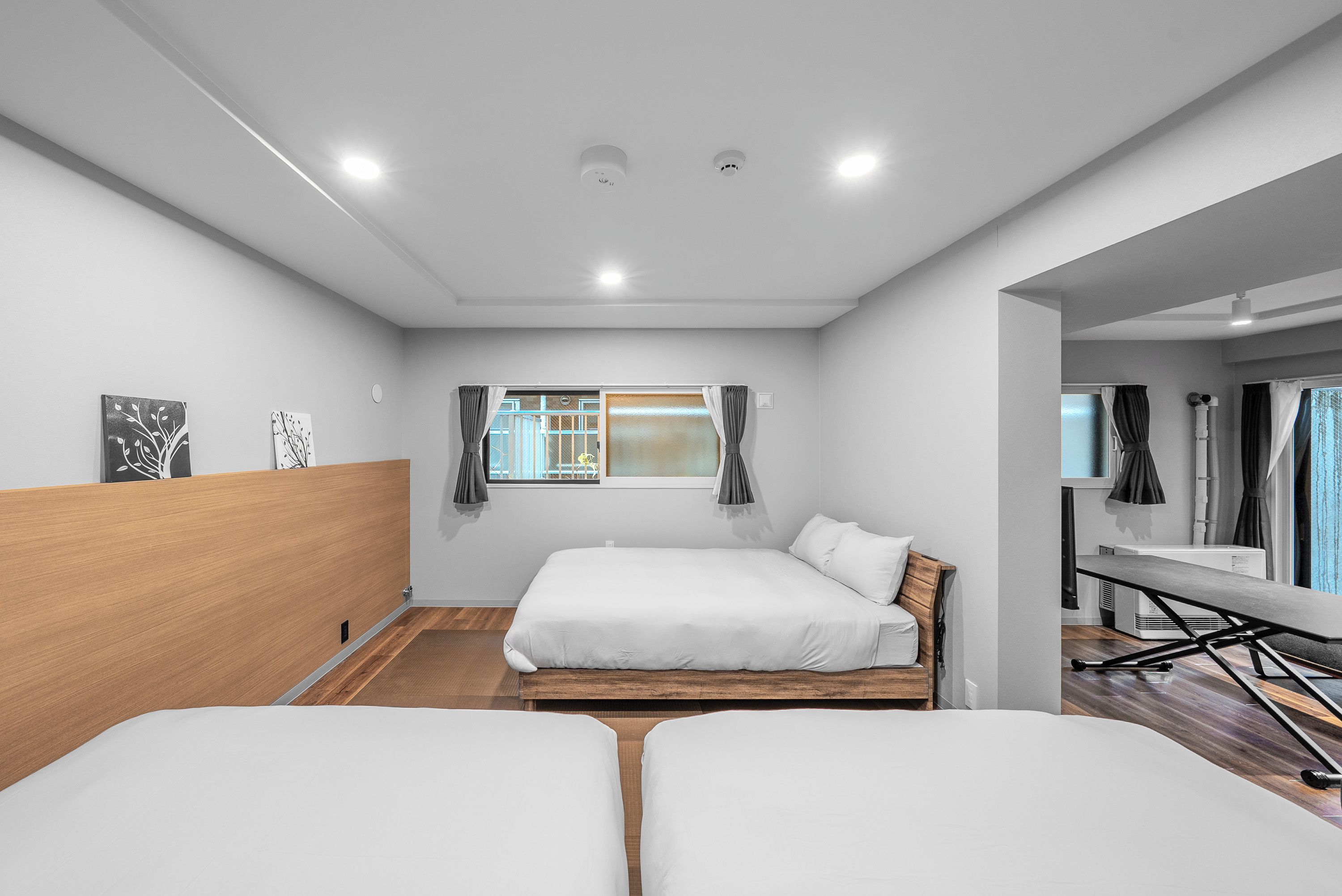 A modern hotel room with two beds, a wooden headboard, a window with curtains, and a small desk with a chair.