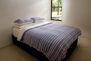 Cama de solteiro com colcha e travesseiros listrados, janela com vista para vegetação e piso coberto de carpete.