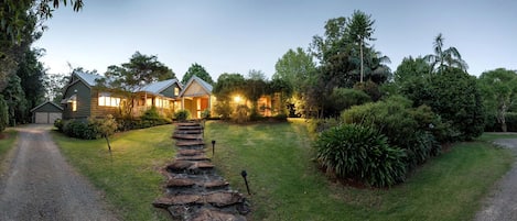 A two-story house with a stone pathway leading to the entrance, surrounded by lush greenery and trees.