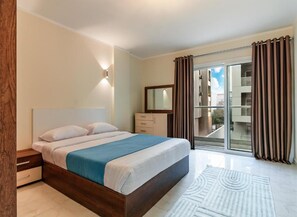 A bedroom with a bed, a nightstand, a dresser, a mirror, and a view through a sliding glass door. - Peaks - El Nessim-1BR-Pool View R106 (Hurghada)