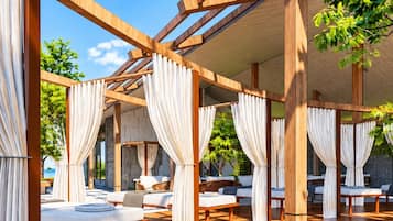 Un espacio al aire libre cubierto con vigas de madera y cortinas blancas, con asientos acolchados y suelo de guijarros.