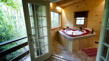 Una cabaña de madera con puerta de vidrio que da a una terraza, un jacuzzi con dos personas y una ventana con vistas a un bosque.