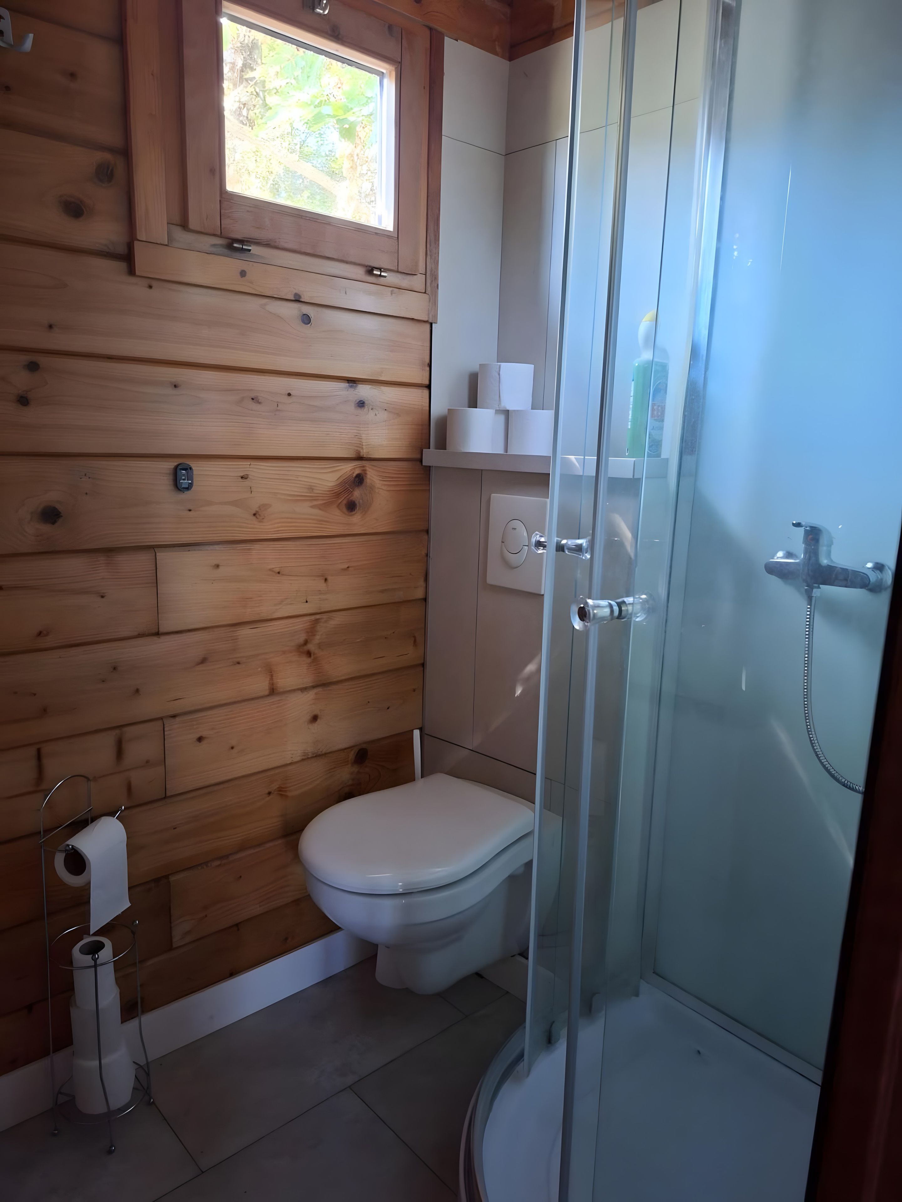 Een badkamer met houten lambrisering, een toilet, een douchecabine van glas en een raam boven de wastafel.