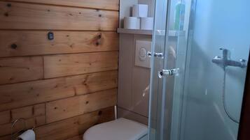 Een badkamer met houten lambrisering, een toilet, een douchecabine van glas en een raam boven de wastafel.