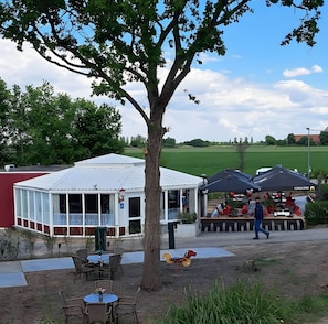 Een terras buiten met tafels en stoelen, een overdekte patio en een grote boom.