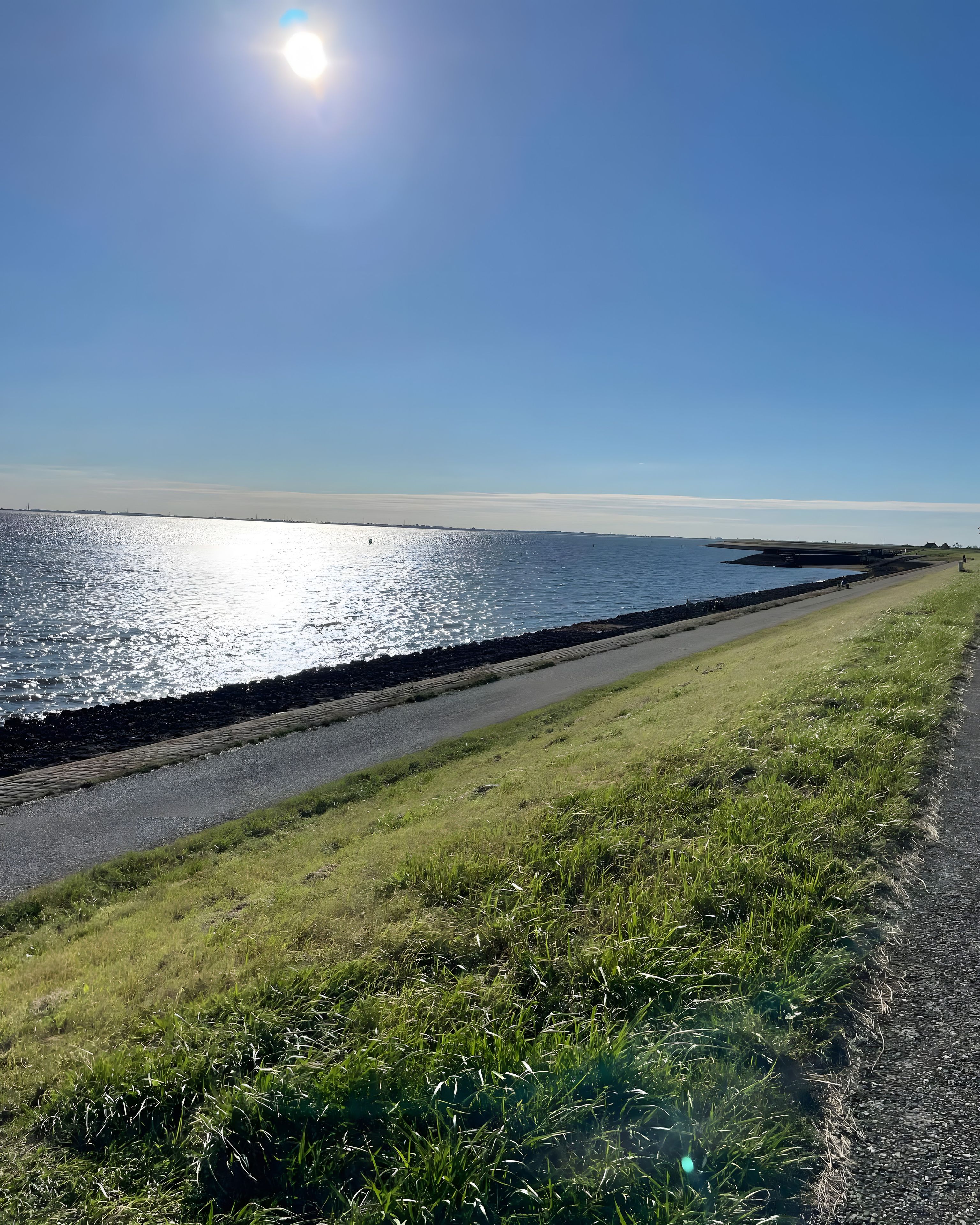 A coastal road with a grassy verge, a paved path, and a body of water reflecting sunlight.