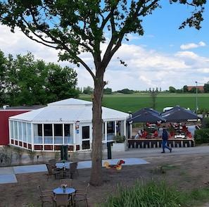 Een overdekt buitengebied met tafels en stoelen, een boom en een gebouw met een wit dak.