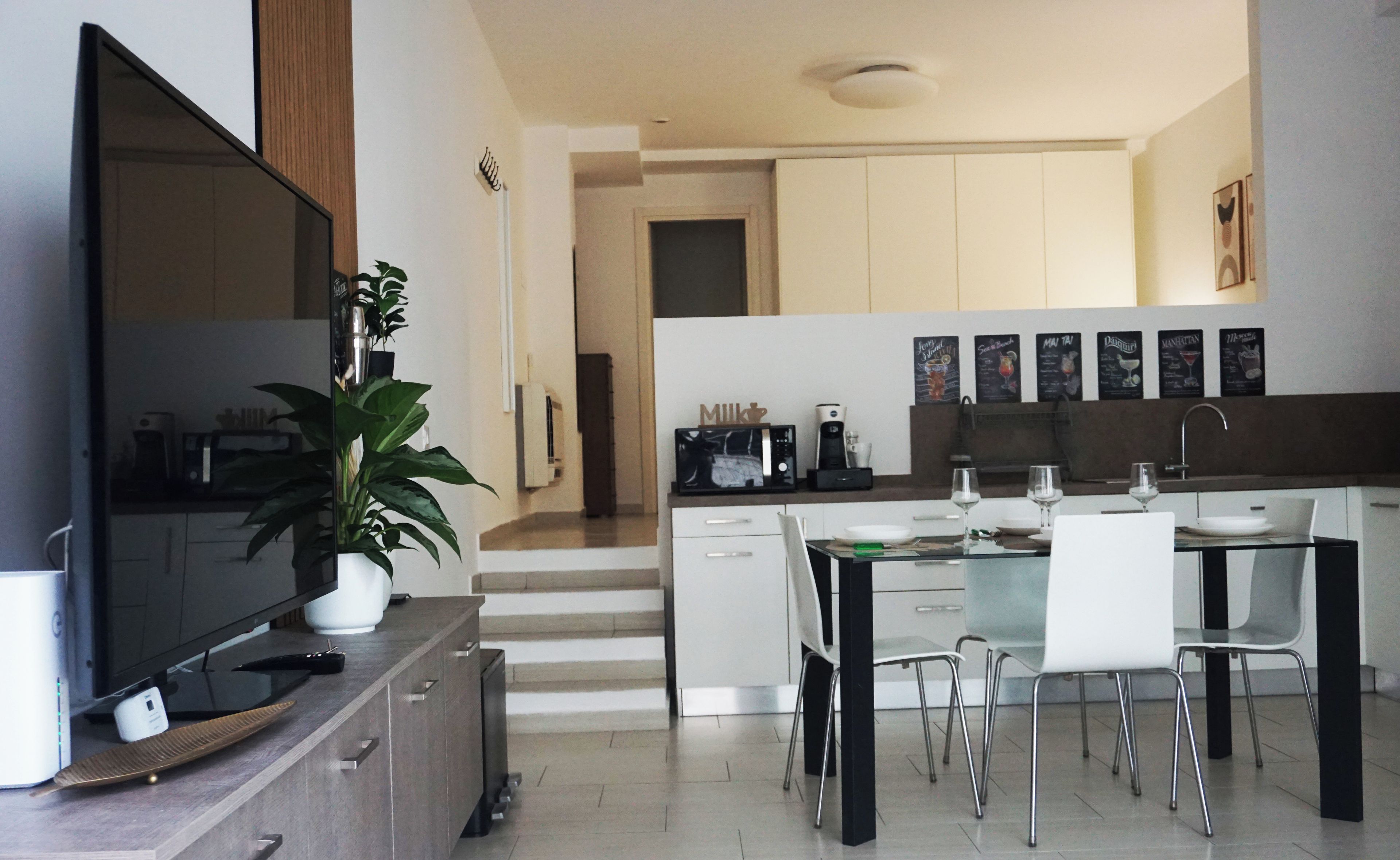 A modern kitchen with a dining area, a flat-screen TV, a coffee maker, and a potted plant.