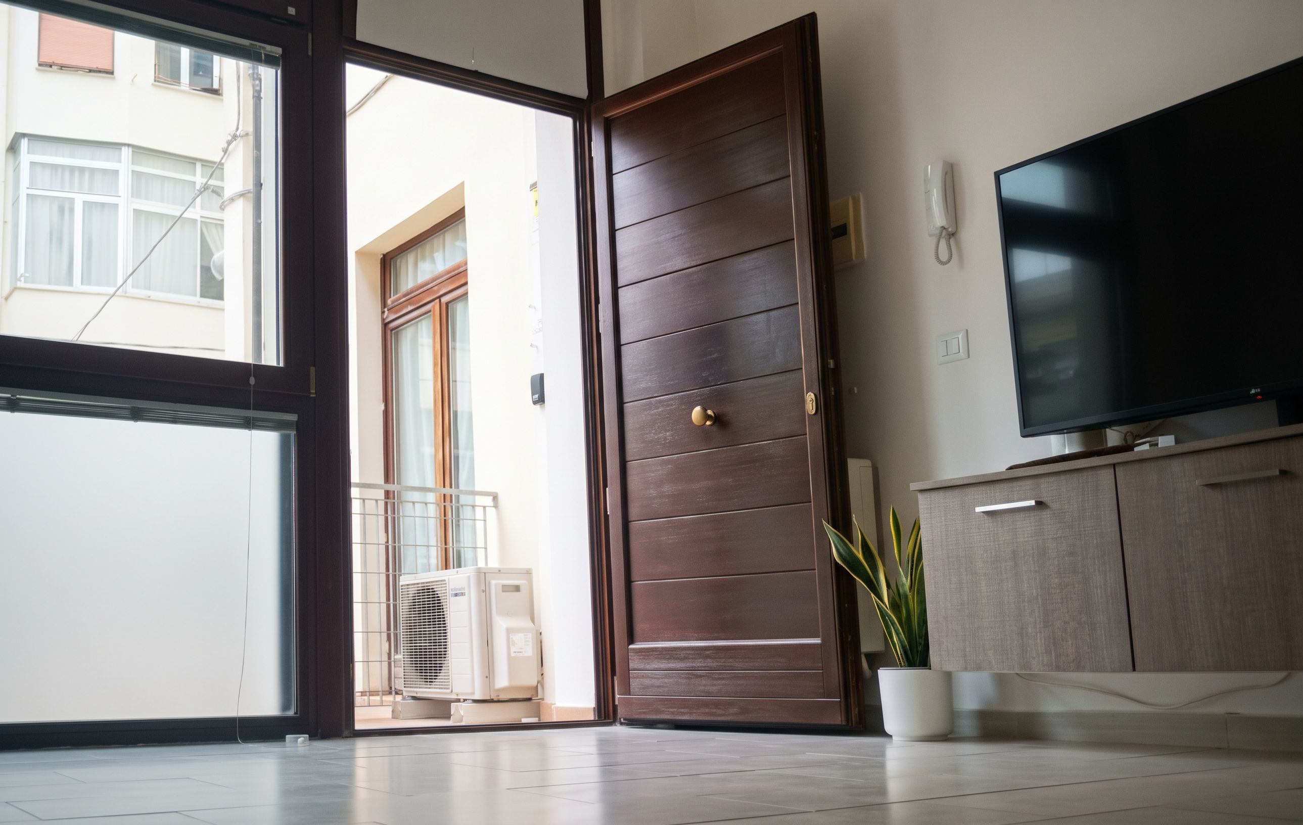 Una habitación con puerta corrediza de vidrio que da a un balcón, un televisor de pantalla plana montado en la pared y una planta en maceta en el suelo.