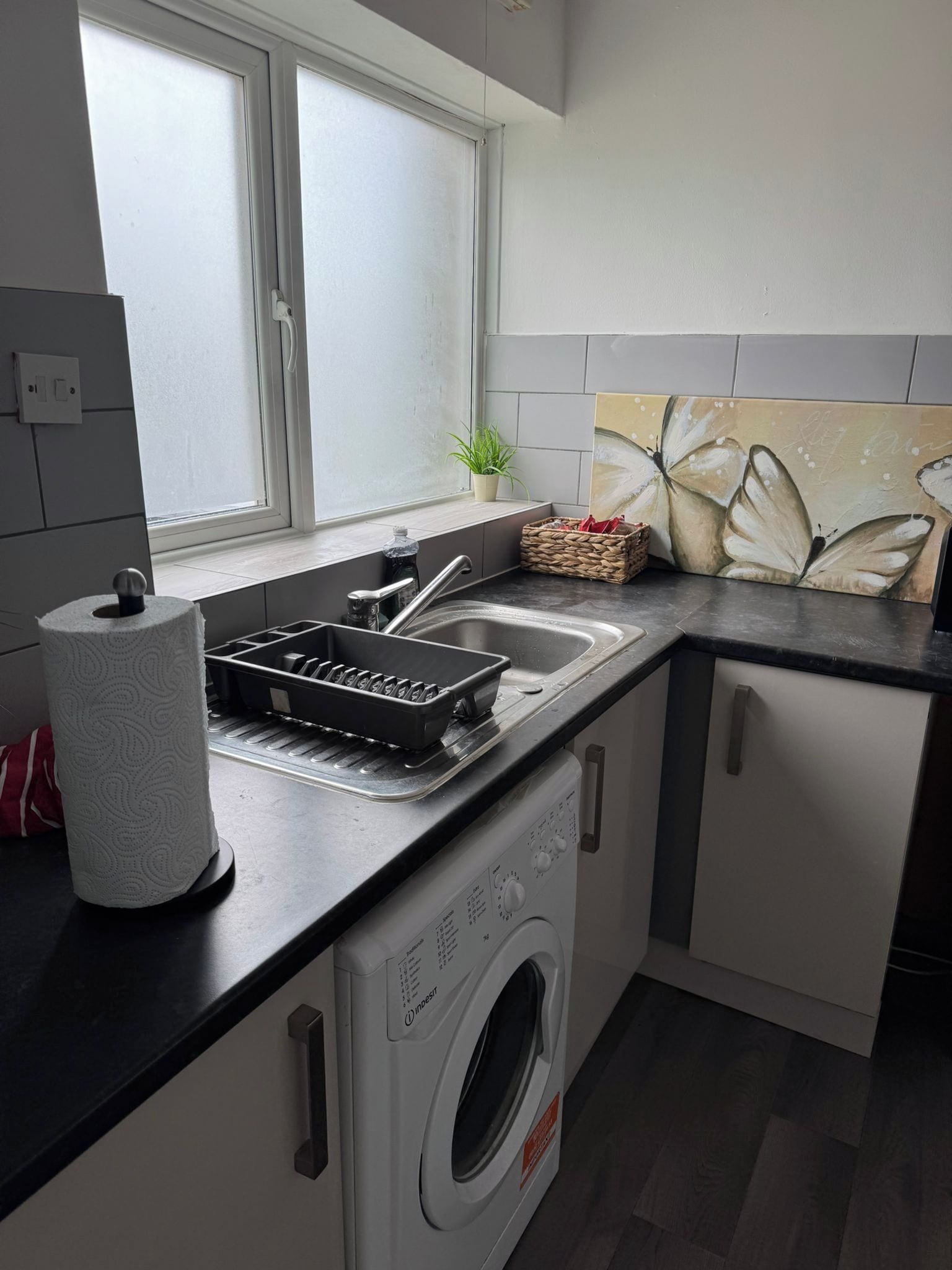 Een kleine keuken met een wasmachine, een spoelbak en een raam met binnengebroken glas.