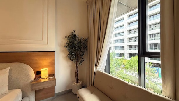 A hotel room with a bed, bedside table with a lamp, a potted plant, a sofa, and a window with curtains. - Maison Sahab - Stylish Studio in Tiraz Building (Sharjah)