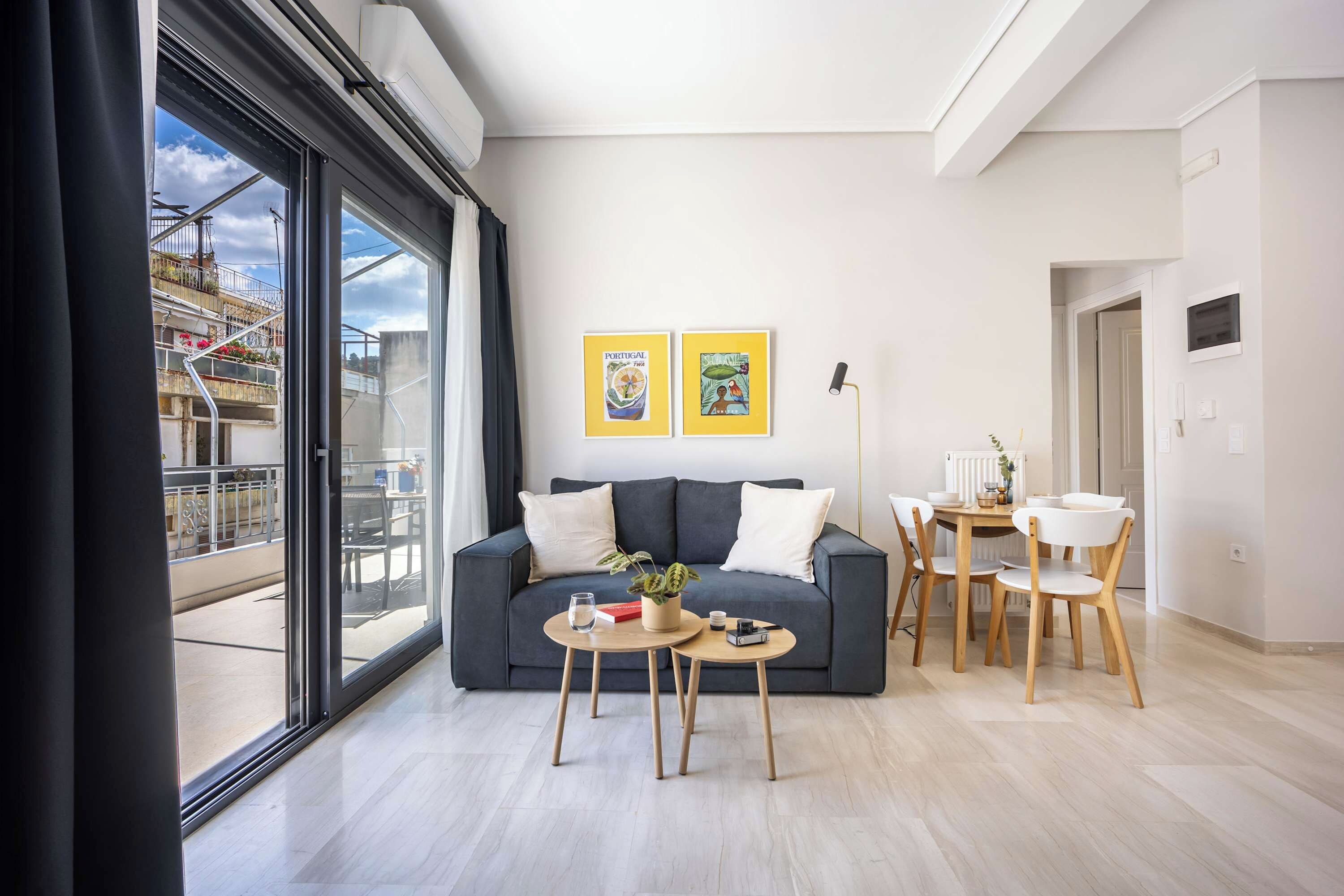 Un salon moderne avec un canapé gris, une table basse et un coin repas avec des chaises en bois. Deux œuvres d’art encadrées ornent le mur, et une grande fenêtre offre une vue sur l’extérieur.