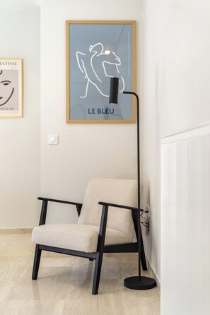A modern chair with a black frame and a light-colored cushion, positioned next to a floor lamp with a black base and a curved neck. - FiftySeven Athens - Two Ways Outside, 2BR | 302 (Athina)