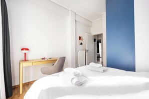 A hotel room with a bed, a desk, a chair, a red lamp, and a door leading to another room. - FiftySeven Athens - 2BR, Terrace & Balcony | 301 (Athina)